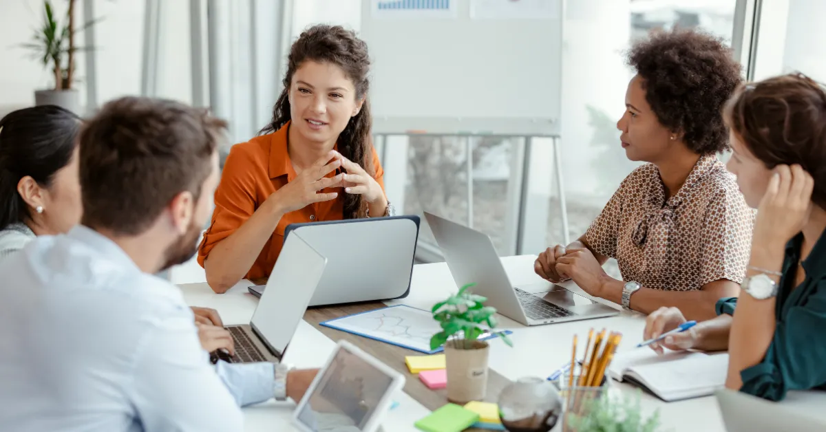 Five colleagues talking around a meeting table with open laptops, a tablet, and notebooks