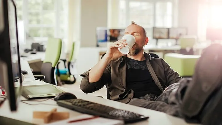 Man relaxing at his office desk, drinking coffee and leaning back comfortably.