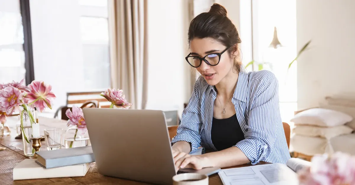 Woman in glasses and a striped shirt typing on a laptop at a home table with pink flowers