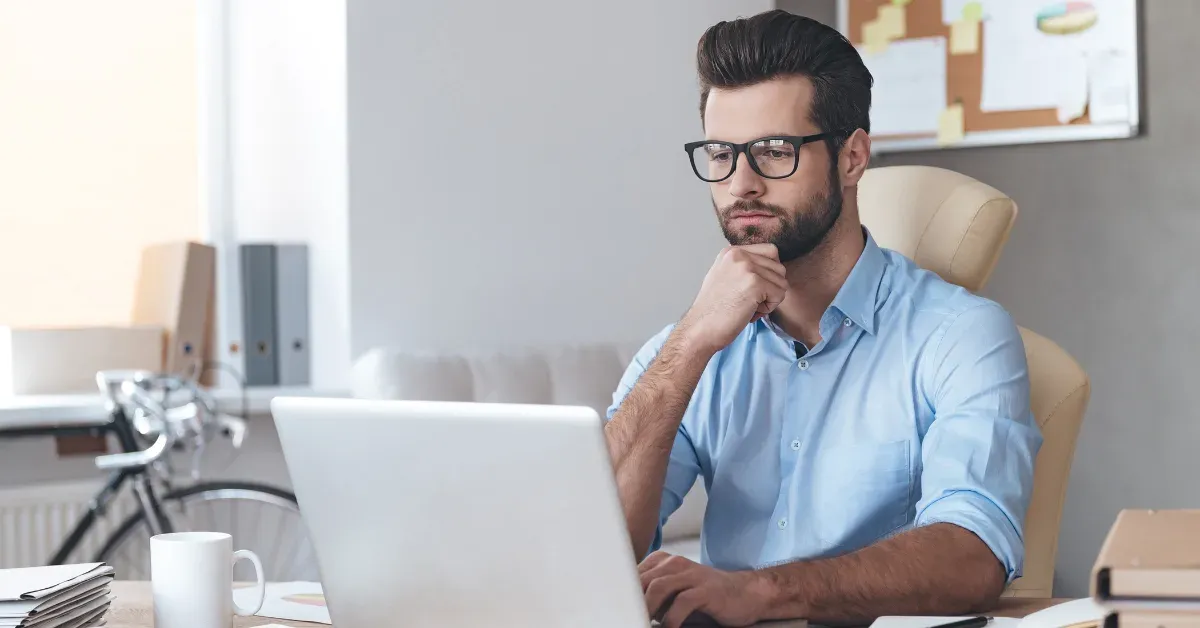 Man in glasses and a light blue shirt resting his chin on his hand while reviewing a laptop at his desk