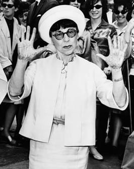 Black-and-white photo of a woman wearing a hat and round glasses, raising both hands, with a crowd in the background.